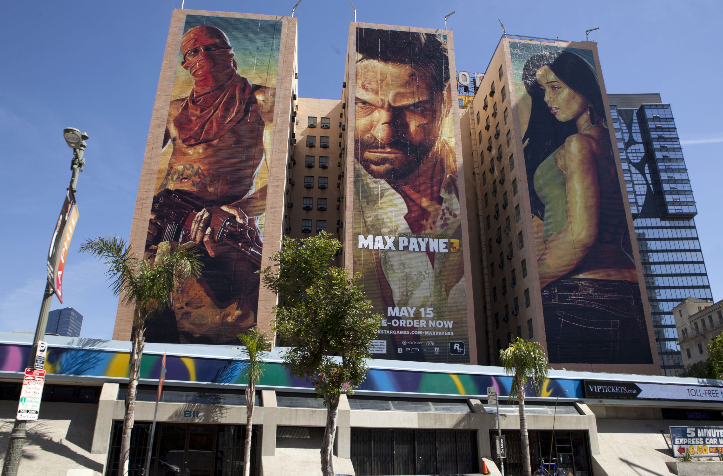 Three tall buildings, each covered with a building-height Max Payne 3 character painting, with palm trees at street level.